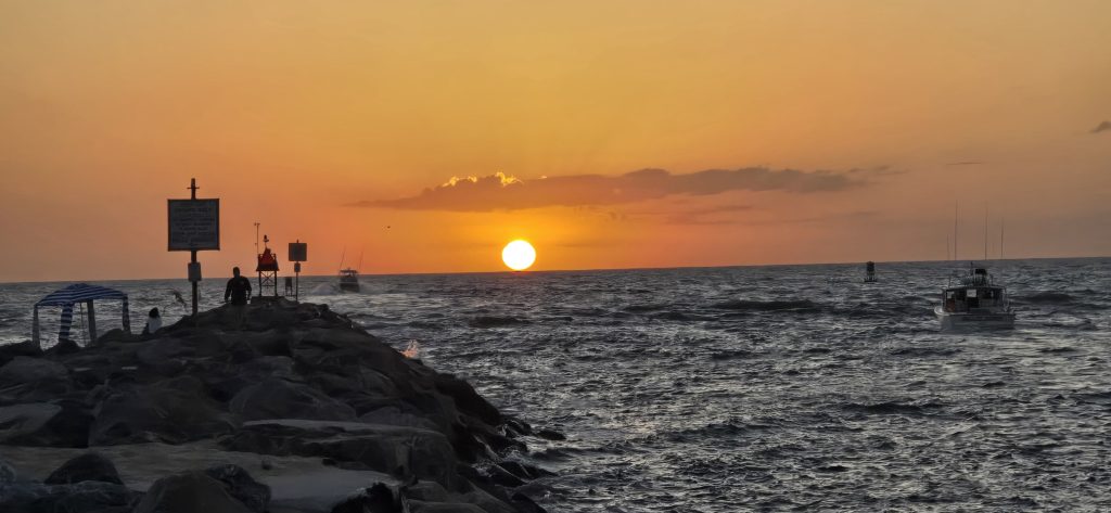 A low orange sun rising over choppy water beyond a rock jetty, with fishing boats and a man standing on the rocks